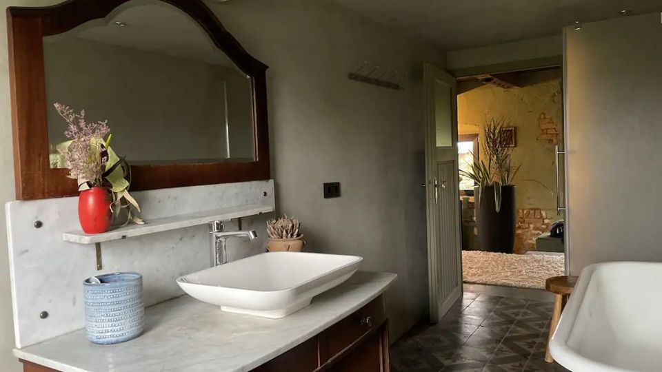 Badezimmer mit einem Waschtisch aus Holz und einer weißen Marmorplatte, darauf ein rechteckiges Keramikwaschbecken mit Armatur. Im Hintergrund ein offener Durchgang zu einem Raum mit Pflanzen und einer gelben Wand.