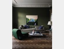 Wohnzimmer mit olivgrüner Wand, moderner grauer Couch, einem grünen Designer-Stuhl und einem niedrigen Couchtisch auf einem strukturierten Teppich. Die Decke hat dekorative Stuckverzierungen, und an der Wand hängt ein großformatiges Kunstwerk mit Naturmotiv.