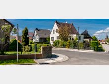 Siedlungsstraße mit freistehenden Einfamilienhäusern, umgeben von Vorgärten mit Bäumen und Sträuchern. Im Vordergrund Gehwege und Straßenschilder, im Hintergrund eine Kirche und weitere Häuser.