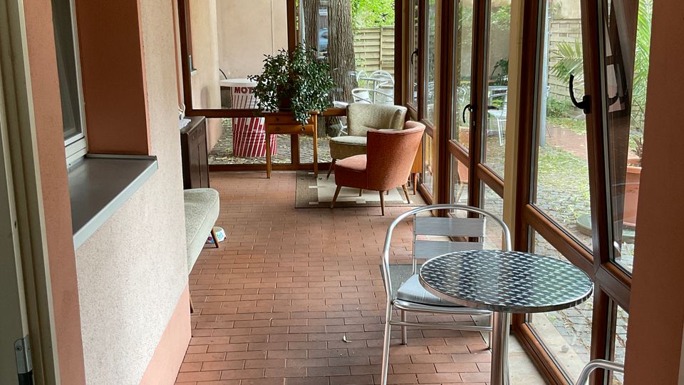 Langer, überdachter Wintergarten mit Ziegelboden, schräger Decke und durchgehender Fensterfront auf der rechten Seite mit Blick in einen kleinen Gartenhof. Im Raum stehen mehrere Sitzgelegenheiten und ein kleiner runder Metalltisch; am Ende befindet sich eine verglaste Tür und weitere Fenster.
