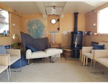 Gemütlicher Innenraum eines Holzhauses oder Schiffs mit großen Fenstern. Zu sehen sind moderne Möbel, darunter ein Sofa, mehrere Sessel, ein Kaminofen und ein Regal mit Büchern, auf hellem Teppichboden.