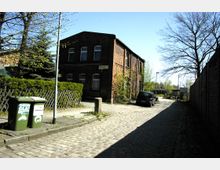 Pflastersteinstraße in einem Wohngebiet mit einem zweistöckigen Backsteingebäude auf der linken Seite, umgeben von einer Hecke und Bäumen. Rechts sind eine Mauer, ein geparktes Auto und im Hintergrund ein kleines Gebäude sichtbar.