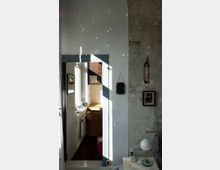 Helles Interieur mit Blick durch eine Türöffnung in einen kleinen Raum mit Fenster und Holzschreibtisch. Auf der rechten Seite ist eine Wand mit abblätterndem Putz, daneben ein Beistelltisch mit Lampe und Telefon; Lichtreflexionen spielen an den Wänden.