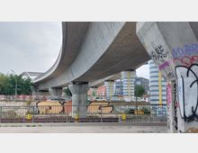 Betonbrücke mit geschwungenem Verlauf, gestützt von Säulen mit blau-weißem Streifenmuster. Im Vordergrund sind ein mit Graffiti besprühter Geländerbereich und weitere urbane Elemente wie Fahrzeuge und Gebäude im Hintergrund sichtbar.