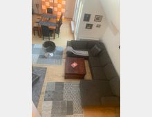 Wohnzimmer mit heller Holzbodenfläche, grauer Eckcouch, einem Holzcouchtisch und gemusterten Teppichen. Im Hintergrund ein Esstisch mit dunklen Stühlen, sowie eine beleuchtete Trennwand und an der Wand Bilderrahmen.