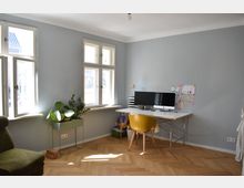 Helles Arbeitszimmer mit Holzfußboden, zwei großen Doppelfenstern, einem Schreibtisch mit Computer und gelbem Stuhl sowie mehreren Zimmerpflanzen in einem Pflanzenständer. Die Wände sind in Pastellgrau gestrichen, und Kinderzeichnungen hängen an der Wand.