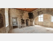 Ein unrenovierter Innenraum mit unverputzten Ziegelwänden, einem gemauerten Rundbogen und mehreren rechteckigen Fenstern, die Tageslicht hereinlassen. Der Raum wirkt roh, mit sichtbaren Schäden und einem offenen Durchgang, der zu einem angrenzenden Bereich führt.