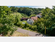 Ländliche Szene mit Hügeln, dichtem Grün und einer kleinen Wohnsiedlung, die aus weißen Häusern mit roten Ziegeldächern besteht. Im Vordergrund führt ein asphaltierter Weg durch die bewachsene Landschaft.