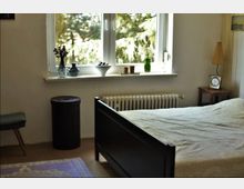 Schlafzimmer mit einem einfachen Bettgestell in dunklem Holz, einem kleinen Nachttisch mit Lampe und Wecker sowie einer Fensterbank mit Dekorationen wie Vasen und Schalen. Das Fenster bietet Ausblick auf grüne Bäume, und unter der Fensterbank befindet sich ein Heizkörper.