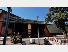 Außenansicht des Theater- und Veranstaltungsgebäudes "Spielbank Berlin" mit großer Glasfassade und moderner Architektur. Im Vordergrund befinden sich Bauarbeiten mit einer Baumaschine, Absperrungen und einer parkenden Motorrad.