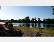 Ein Uferbereich mit grünem Rasen, mehreren roten Liegestühlen auf einer Holzterrasse und einer Metalltreppe, die ins Wasser führt. Im Hintergrund ein ruhiger Fluss, gesäumt von Bäumen und Vegetation, unter einem klaren, blauen Himmel.