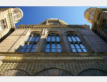 Fassadenansicht einer historisch gestalteten Synagoge mit reich verzierten Ornamenten, großen Rundbogenfenstern und hebräischer Inschrift über dem Eingang. Zwei seitliche Kuppeltürme und der zentrale Hauptturm ragen in den klaren, blauen Himmel.
