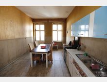 Schmale Küche in einem älteren Gebäude mit gelben Wänden und hohen Decken. Links steht ein Esstisch mit Rattanstühlen, rechts eine Küchenzeile mit weiß-blauen Schränken, Spüle und Herd, am Ende des Raumes zwei große Fenster mit Rundbögen.