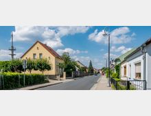 Straßenszene in einem Wohngebiet mit Einfamilienhäusern, Vorgärten und Laternen entlang eines Gehwegs. Im Hintergrund ist ein Telekommunikationsturm sichtbar, begleitet von einer lockeren Bebauung und einem blauen Himmel mit weißen Wolken.
