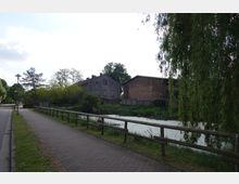 Fußweg entlang eines kleinen Kanals mit hölzernem Geländer, umgeben von Grünflächen und Bäumen. Im Hintergrund stehen zwei ältere Gebäude, eines aus grauem Putz und eines aus Backstein, umgeben von Vegetation.