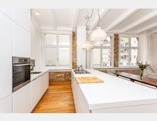 Helle, moderne Wohnküche mit weißen Einbauten und großer Kücheninsel, Holzfußboden und sichtbaren Backsteinpfeilern. Große Fensterfronten und zwei hängende Glaslampen beleuchten den Raum; daneben steht ein Esstisch am Fenster.