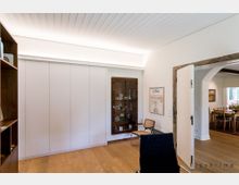 Heller Wohnraum mit Holzboden und Einbauschrank mit weißen Türen. Im Hintergrund ist ein Glastürenschrank mit dekorativen Gegenständen, ein Holzrahmen trennt den Raum von einem angrenzenden Essbereich mit Holzmöbeln.