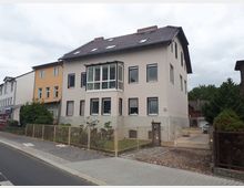 Mehrstöckiges Wohnhaus mit hell verputzter Fassade, Satteldach mit Dachfenstern und einem verglasten Erker an der Straßenseite. Vor dem Gebäude befinden sich ein kleiner, eingezäunter Vorgarten sowie eine Einfahrt mit Tor und gepflastertem Bereich entlang eines Gehwegs.