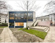 Zweistöckiges modernes Gebäude mit dunkelblauer Fensterfassade und großen Glasflächen, gelegen in einem Innenhof mit Rasenflächen und kahlen Bäumen. Rechts führt eine breite Außentreppe aus Beton zu einer erhöhten Plattform neben einer niedrigen Mauer.