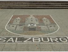 Ein Mosaik auf einem gepflasterten Platz zeigt das Wappen von Salzburg mit einer stilisierten Burg und der Schrift "SALZBURG" darunter. Im Hintergrund sind breite Steinstufen erkennbar.
