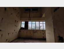 Ein leerer, verfallener Raum mit abgeblättertem Putz an den Wänden und einem großen, geteilten Fenster. Durch das Fenster sind schemenhaft Außenstrukturen wie Strommasten erkennbar.