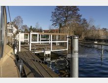Eine hölzerne Uferpromenade mit einem Geländer und einer angrenzenden Plattform am Wasser, umgeben von Schilfpflanzen und Bäumen im Hintergrund. Im Vordergrund sind Festmacherleinen und Geländerfunde, während im Hintergrund Wohnhäuser und ein klarer Himmel sichtbar sind.