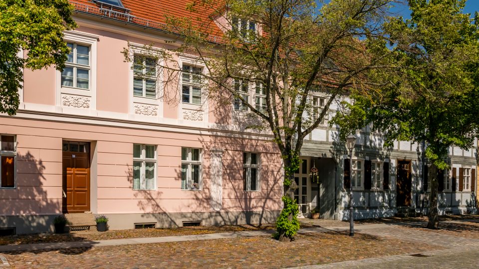 Straßenansicht einer Reihe historischer Wohnhäuser mit pastellfarbenen Fassaden, weißen Fensterrahmen und roten Ziegeldächern. Davor verläuft eine gepflasterte Straße mit Gehweg und mehreren großen Bäumen, die Schatten auf die Fassaden werfen.
