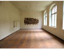 Ein leerer Raum mit heller, leicht verwitterter Wandgestaltung und einem Parkettboden in Fischgrätmuster. Rechts befinden sich drei große, bogenförmige Fenster, die Tageslicht einlassen, während an der Stirnwand ein kunstvolles Wandrelief angebracht ist.