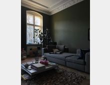 Wohnzimmer mit dunkelgrüner Wand und verziertem Stuck an der Decke. Ein graues Sofa, ein niedriger Couchtisch mit Büchern und Dekoration sowie ein großer Fensterbogen mit Tageslicht und einer Zimmerpflanze prägen den Raum; auf dem Boden liegt ein strukturiert wirkender Teppich.