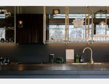 Moderne Küchenzeile mit dunkler Arbeitsplatte, hellgoldenem Wasserhahn und gläsernen Regalen mit Geschirr und Gläsern. Im Hintergrund sind große Fenster mit Blick auf Bäume und städtische Gebäude erkennbar.