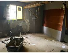 Ein verlassener, heruntergekommener Raum mit beschädigter Wandverkleidung, einem zerbrochenen Fenster mit teilweisem Vorhang und herumliegenden Kabeln. Auf dem Boden liegen Schutt und eine alte Plastikwanne, während mehrere alte Rohrleitungen und Metallhaken an der Decke angebracht sind.