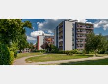 Mehrstöckige Wohngebäude mit Balkonen, umgeben von Bäumen und gepflegten Grünflächen. Ein geschwungener Gehweg führt durch die begrünte Umgebung, der Himmel ist teilweise bewölkt.