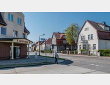 Straßenszene in einem Wohngebiet mit einer Apotheke an der Ecke eines Gebäudes, das weiß-rote Fassaden hat, und mehreren Einfamilienhäusern mit roten Ziegeldächern entlang der Straße. Ein Gehweg, Straßenlaternen und ein Fußgängerüberweg sind ebenfalls sichtbar.