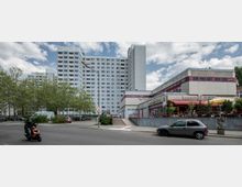 Mehrstöckiges Wohngebäude in moderner Plattenbauweise im Hintergrund, flankiert von mehreren Bäumen. Im Vordergrund befindet sich ein Flachbau mit einem Restaurant, vor dem Sitzgelegenheiten und Sonnenschirme stehen; auf der Straße fahren ein Auto und ein Motorroller.