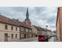 Straßenszene in einer kleinen Stadt mit niedrigen Wohnhäusern und roten Ziegeldächern, im Hintergrund ein Kirchturm mit dunkler Spitze. Am Straßenrand parken Autos, darunter ein roter Lieferwagen, und die Straße ist mit Kopfsteinpflaster gepflastert.