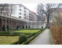 Ein symmetrisch angelegter Garten mit geschnittenen Hecken, Rasenflächen und kleineren Buchsbaumkugeln, flankiert von einer Mauer mit Arkaden. Im Hintergrund sind mehrstöckige Wohngebäude und laublose Bäume zu sehen.