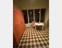 Raum mit einem karierten Boden in Braun- und Beigetönen, einem großen, roten Holzschrank links, einem runden weißen Tisch mit zwei Holzstühlen rechts und einem großen Fenster im Hintergrund, durch das eine dunkle, beleuchtete Innenszene zu sehen ist.