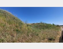 Gräserbewachsene Hügel unter klarem, blauem Himmel, mit einem schmalen, unbefestigten Weg auf der rechten Seite des Bildes. Im Hintergrund sind vereinzelt Büsche und Bäume zu sehen.