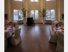 Großer, heller Raum mit hohen Decken und Holzboden, eingerichtet mit runden Tischen, die mit weißen Tischdecken und Stuhlbezügen gedeckt sind. Im Hintergrund steht ein schwarzes Klavier vor großen Fenstern mit Blick auf eine Außenlandschaft, flankiert von hohen Pflanzen in weißen Vasen.