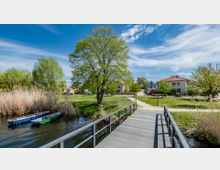 Ein Park im Frühling mit einem Holzsteg über ein Gewässer, an dessen Ufer Schilf und kleine Boote liegen. Im Hintergrund sind Wiesen, Bäume, Fußwege und Wohnhäuser zu sehen, teilweise mit Menschen auf Fahrrädern.
