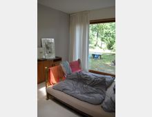 Ein Schlafzimmer mit einem niedrigen Bett, bedeckt mit grauer Bettwäsche und farbigen Decken. Im Hintergrund befindet sich ein großes Fenster mit Blick auf einen grünen Garten, neben dem ein Vorhang hängt. Links steht eine Holzkommode mit darauf abgestellten Bildern.