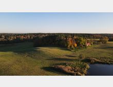 Landschaft mit sanften Wiesenhügeln, einem kleinen Teich im Vordergrund und einer Baumgruppe mit herbstlich gefärbten Blättern, hinter der sich ein einzelnes Haus mit rotem Dach befindet; im Hintergrund ein dichter Wald unter klarem Himmel.