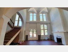 Großer, heller Raum mit hohen Decken und großen Spitzbogenfenstern. Eine Holztreppe führt nach oben, während sich unten ein Kamin und eine Tür mit verzierten Glas- und Gitterelementen befinden.