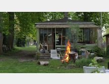 Kleines, einfaches Gartenhaus mit verglaster Veranda, umgeben von Bäumen und grünem Gras. Im Vordergrund brennt ein offenes Feuer in einer Feuerstelle, umgeben von Gartenstühlen und Holzstücken.