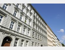 Mehrstöckige, hellgraue Wohngebäude im historischen Stil mit symmetrischen Fassaden, dekorativen Fensterrahmen und Gesimsen, rechts daneben weitere ähnliche Gebäude. Der Himmel ist klar und blau.