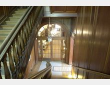 Holzvertäfeltes Treppenhaus mit einer gewundenen Holztreppe und Balustrade, die zu einer Glastür mit Holzrahmen und Oberlicht führt. Im Hintergrund ist ein Kronleuchter sichtbar, der einen beleuchteten Raum andeutet.