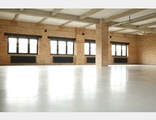 Großer, leerer Raum mit hellen Ziegelwänden, mehreren großen schwarzen Fenstern und Sichtbetondecke. Der Fußboden ist glatt und hell, Heizkörper befinden sich unter den Fenstern.