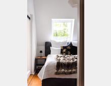 Schlafzimmer mit einem Bett, das mit weißen Kissen, einem schwarzen Dekokissen mit goldenen Blättern und einer braunen Kunstfelldecke dekoriert ist. Links neben dem Bett steht ein kleiner Nachttisch mit einer schwarzen Lampe, und im Hintergrund befindet sich ein Dachfenster mit Blick ins Grüne.