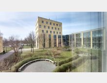 Ansicht eines modernen Campusgebäudes mit gelber Klinkerfassade und großen Fenstern. Im Vordergrund eine gepflegte Grünanlage mit kleinen Wegen, Bänken und Bäumen; im Hintergrund sind angrenzende Bürogebäude mit Glasfronten sichtbar.