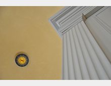 Detailansicht einer Innenraumdecke mit hellgelber Wand und einer blauen Rosette in der Mitte, verziert mit einer goldenen Sternform. Rechts im Bild ist ein Abschnitt einer weißen, klassizistischen Säule mit gerilltem Design erkennbar.